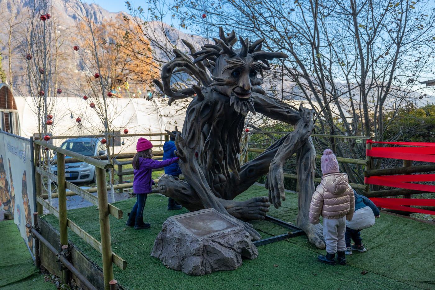 Bambini che esplorano la scultura dell’albero vivente nel Bosco dei Twergi alla Grotta di Babbo Natale, con le montagne di Ornavasso