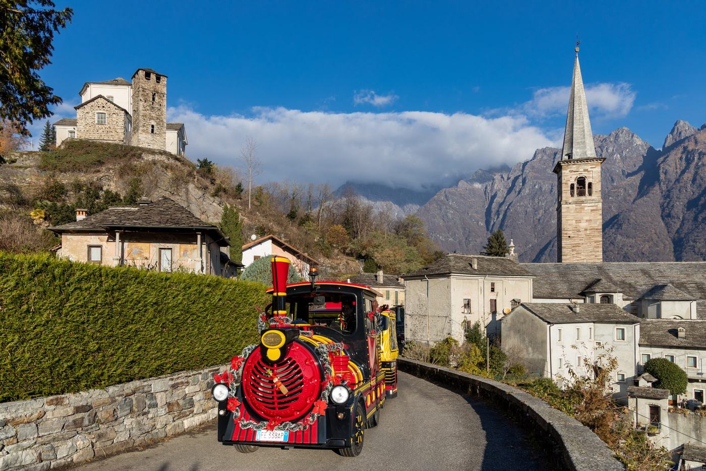 Trenino turistico natalizio Renna Express che attraversa il borgo di montagna di Ornavasso, con campanile e Alpi sullo sfondo
