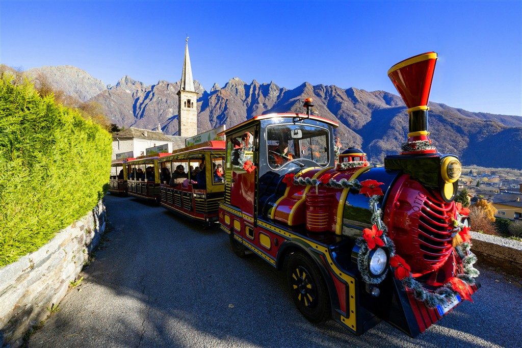 Trenino Renna Express della Grotta di Babbo Natale: locomotive rossa e carrozze gialle sul sentiero di montagna verso la Chiesa della Guardia a Ornavasso