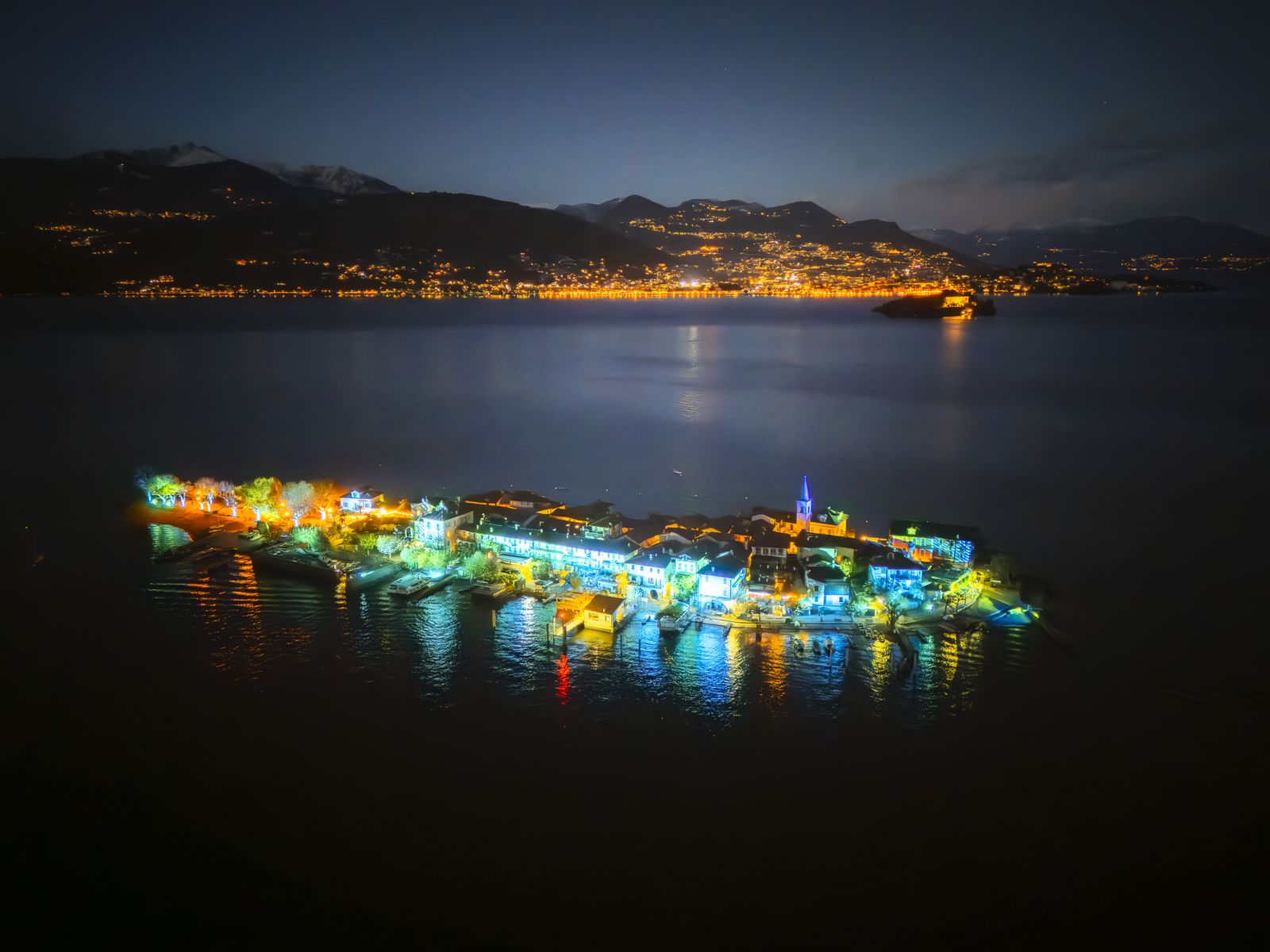Veduta aerea notturna dell’Isola Pescatori illuminata di ciano e oro per le Isole di Luce, con la costa di Baveno sullo sfondo