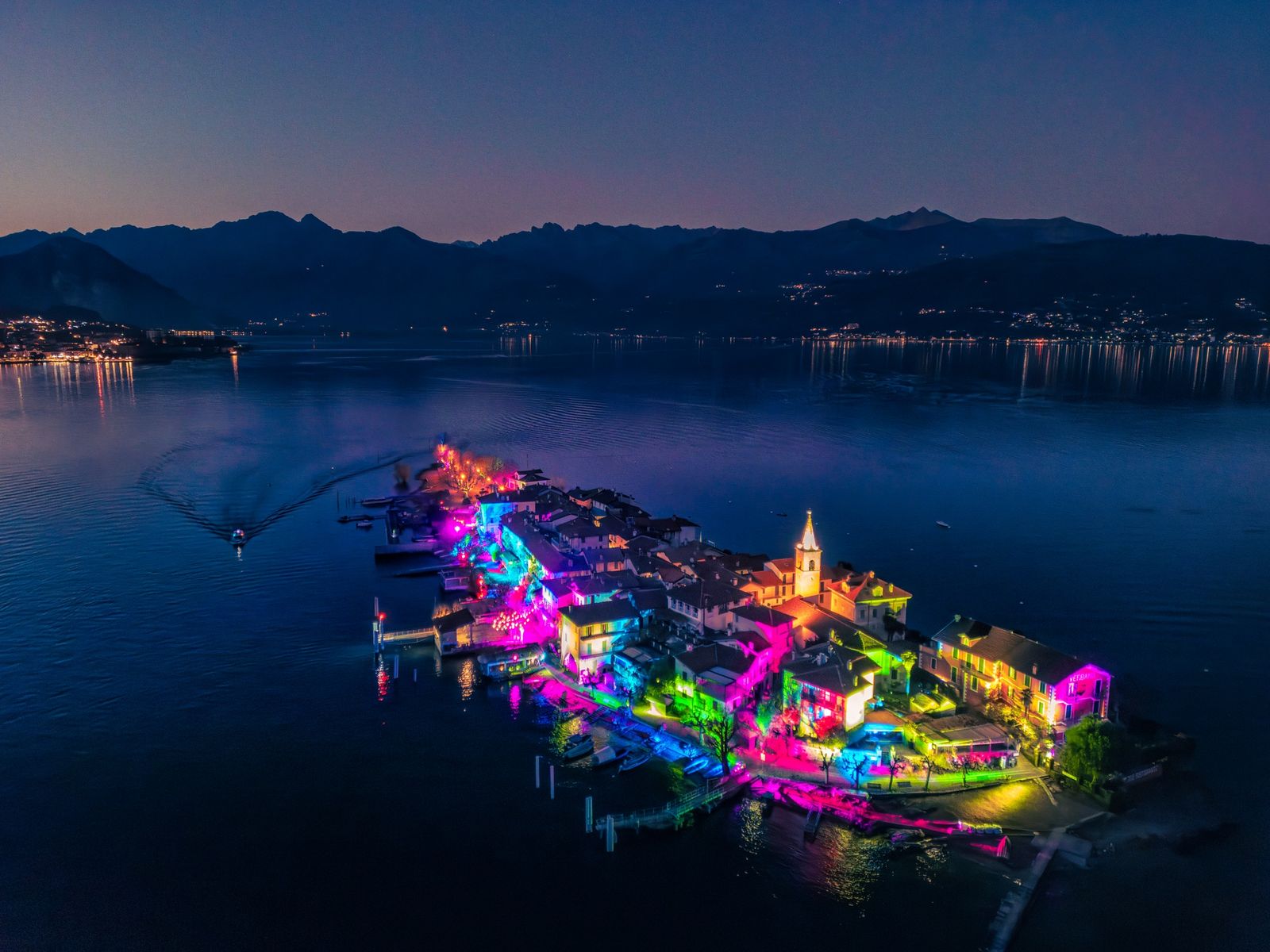 Isola Pescatori illuminata di magenta e blu per le Isole di Luce, con motoscafo in avvicinamento sul Lago Maggiore
